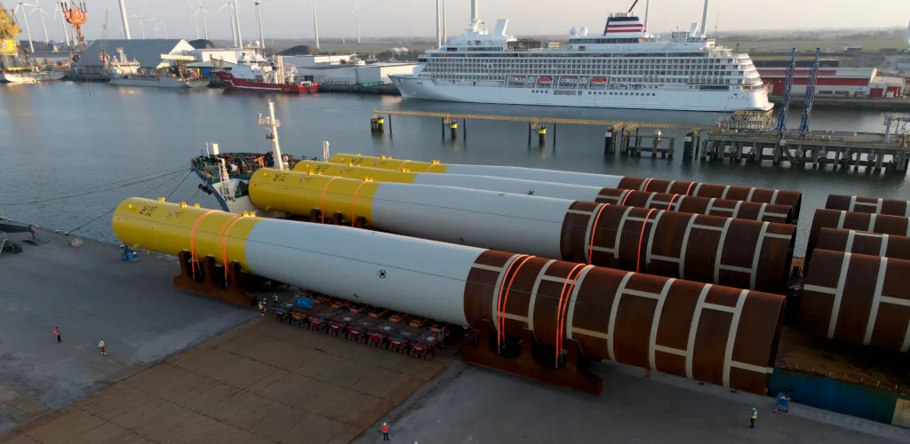 Offshore wind monopile foundations being handled at a port terminal beside docked vessels.
