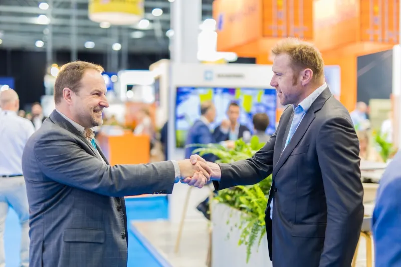 Two Breakbulk Europe attendees shake hands while networking on the busy exhibition floor.